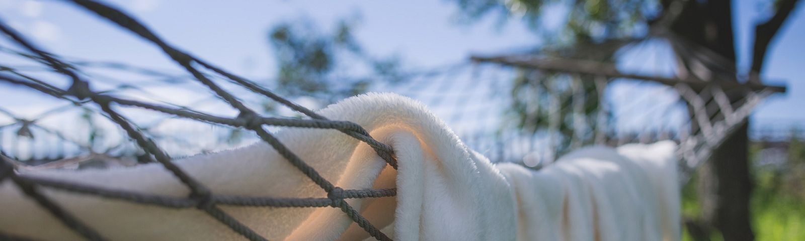 1710-Ontspannen-maar-hoe-dan-1600×480 Weißes, flauschiges Handtuch in einer Hängematte, im Hintergrund verschwommene Bäume und blauer Himmel – ideal für Sportler.