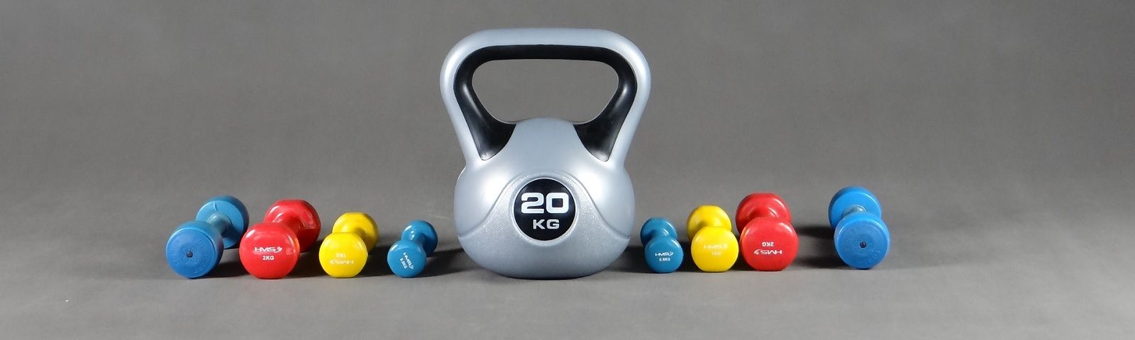 Ein großer silberner Kettlebell mit 20 kg Gewicht, umgeben von bunten Kurzhanteln in Rot, Gelb und Blau.