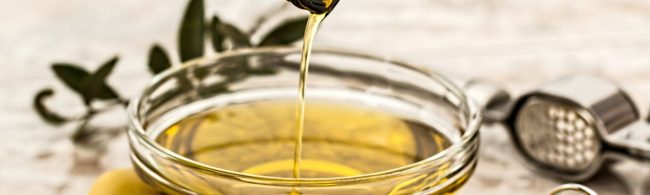 Olivenöl fließt in eine Schüssel, mit Olivenzweigen und Knoblauchpresse im Hintergrund.