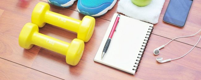 Gelbe Kurzhanteln, Trainingsschuhe, Notizbuch, Stift, Kopfhörer, Handy, Apfel auf Handtuch.
