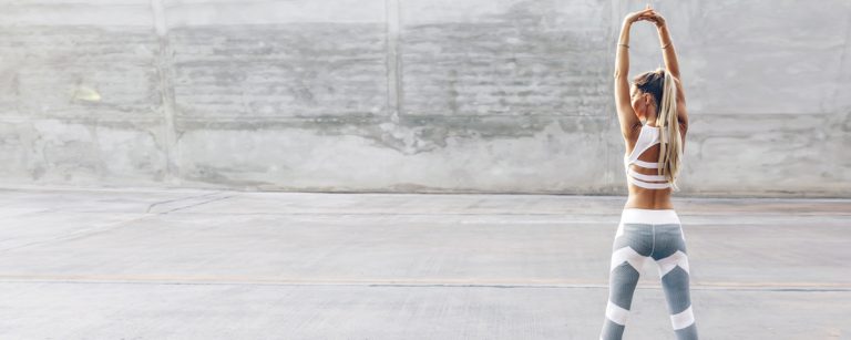 Sportlerin in weißem Sport-BH und grau-weißer Leggings streckt Arme über dem Kopf, Fitness und gesunde Ernährung.