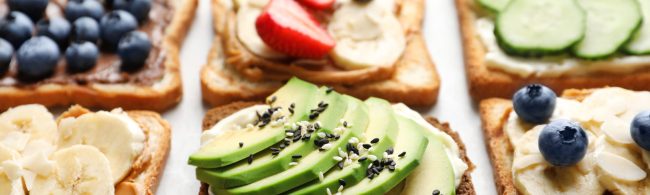 A tray with three healthy sandwich options: a banana‑topped nut butter slice, avocado power toast with seeds, and a Greek‑style sandwich with tzatziki, feta, cucumber, tomato and herbs.