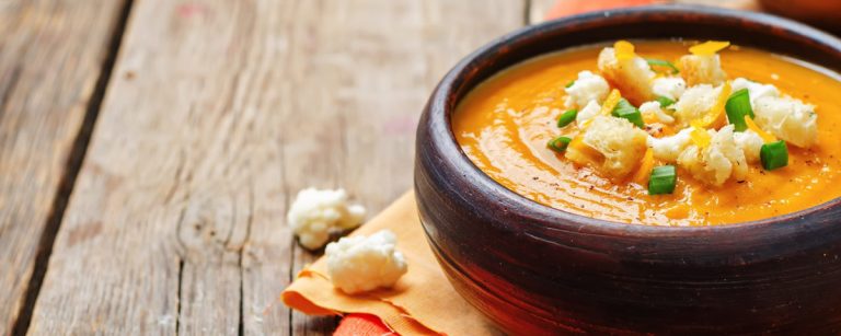 A steaming bowl of creamy sweet potato and cauliflower soup, garnished with fresh parsley, placed on a wooden table.