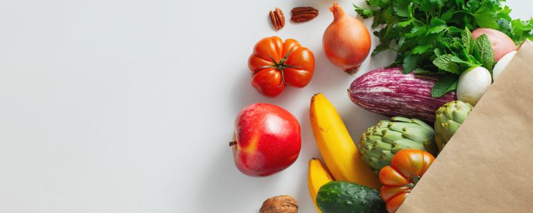 aper grocery bag with assorted seasonal produce—tomatoes, apple, bananas, eggplant, cucumber, artichokes, onion, fresh herbs and nuts—on a light background