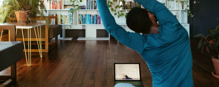 Eine Person macht eine Yoga-Dehnung zu Hause auf einer Matte, folgt einem Online-Fitnesskurs auf einem Laptop. Heimtraining für Flexibilität, Gesundheit und Wohlbefinden in einem inspirierenden Umfeld mit Pflanzen und Büchern.