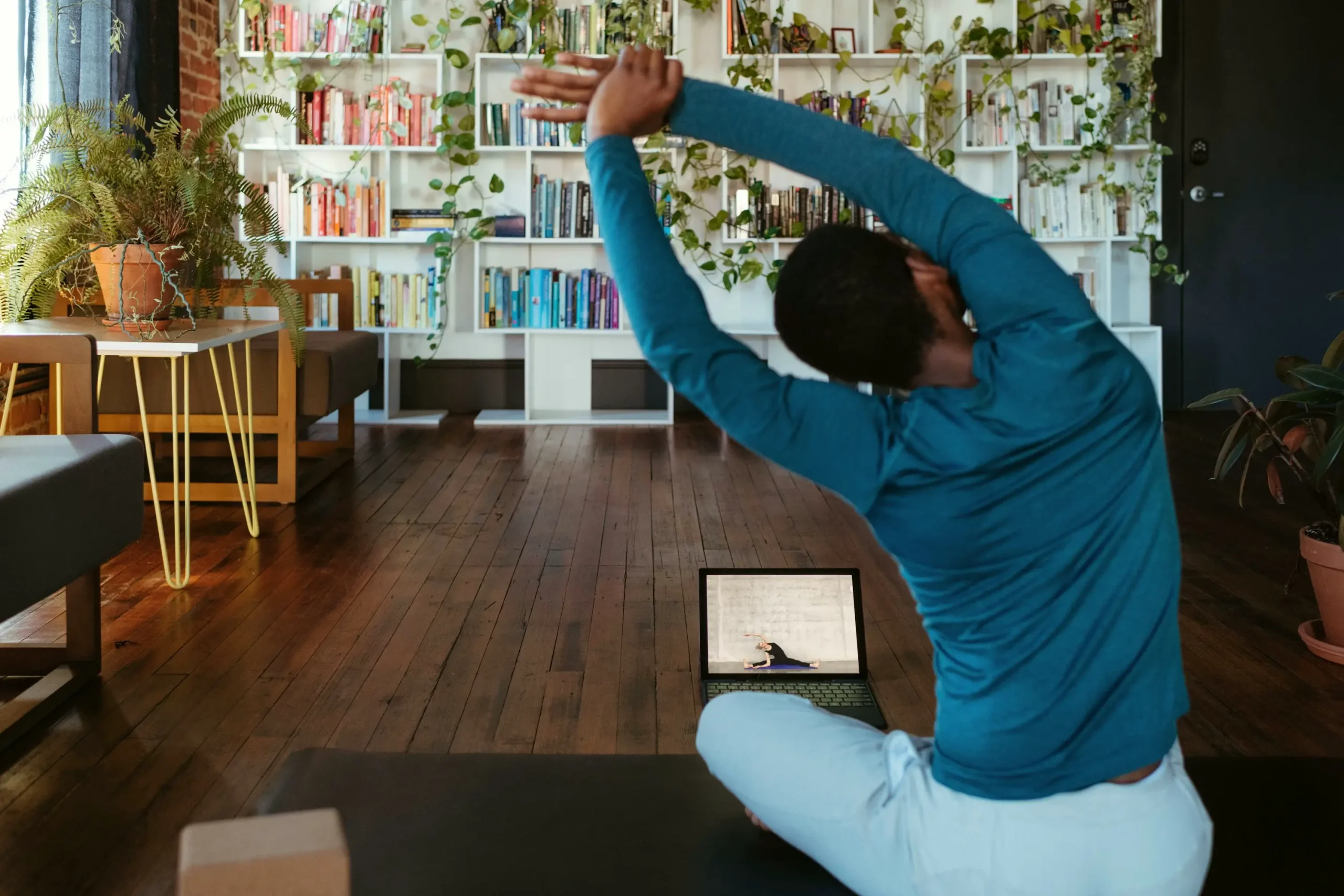 online-fitness-coach-2025 Eine Person macht eine Yoga-Dehnung zu Hause auf einer Matte, folgt einem Online-Fitnesskurs auf einem Laptop. Heimtraining für Flexibilität, Gesundheit und Wohlbefinden in einem inspirierenden Umfeld mit Pflanzen und Büchern.