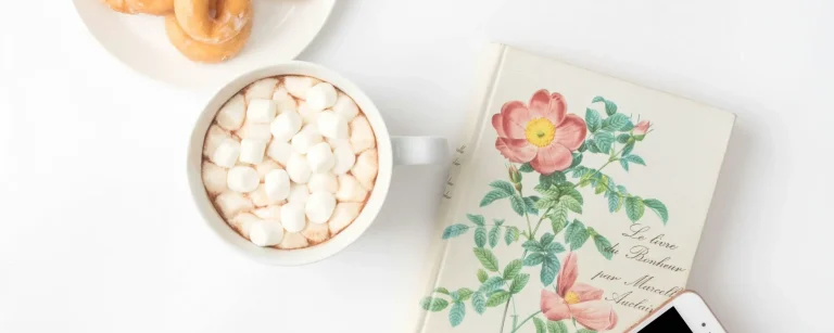 Draufsicht auf eine gemütliche Szene mit glasierten Donuts, Kakao mit Marshmallows, einem floralen Buch und einem Smartphone auf weißem Hintergrund. Dieses Bild kann einen Moment der Entspannung und des Genusses symbolisieren, um die Wichtigkeit von **Balance** und realistischer Integration von Leckereien in einen **gesunden Lebensstil** und eine **Fitness-Ernährung** zu unterstreichen.